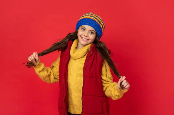 Niño positivo con pelo rizado en sombrero de invierno de punto y chaleco abajo sobre fondo rojo, mantener caliente —  Fotos de Stock