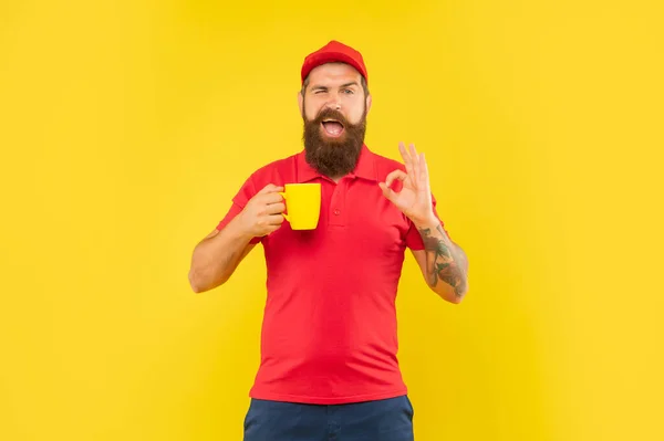 Felice ammiccante ragazzo in casual cap rosso e tshirt dando OK tenendo tazza sfondo giallo, caffè — Foto Stock