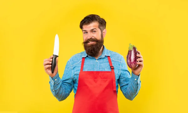 Happy man in apron holding chefs knife and eggplant yellow background, chef — Stock Photo, Image