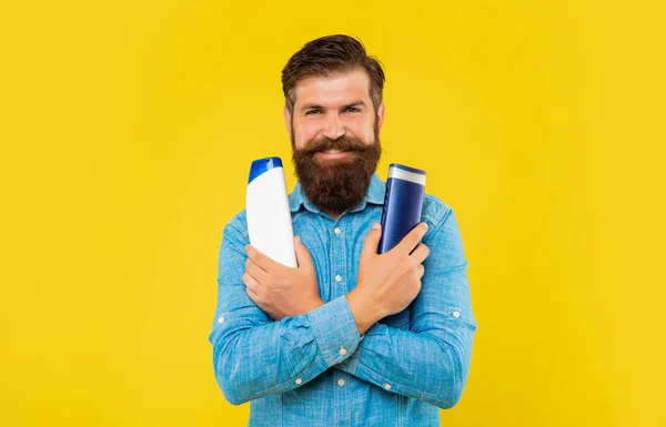 Улыбающийся жестокий бородатый мужчина выбирает бутылку с кондиционером для волос, представляет туалетные принадлежности. — стоковое фото