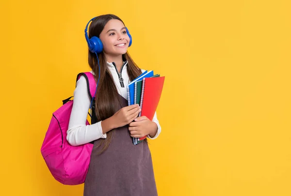 Zadowolone dziecko w słuchawkach ze szkolnym plecakiem z skoroszytem na żółtym tle — Zdjęcie stockowe