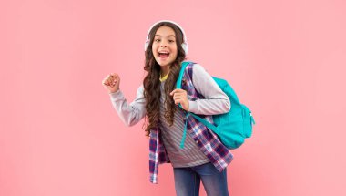 Kulaklıkla müzik dinleyen, okul çantalı, pembe arka planda, çocukluğunda hayrete düşmüş bir çocuk.