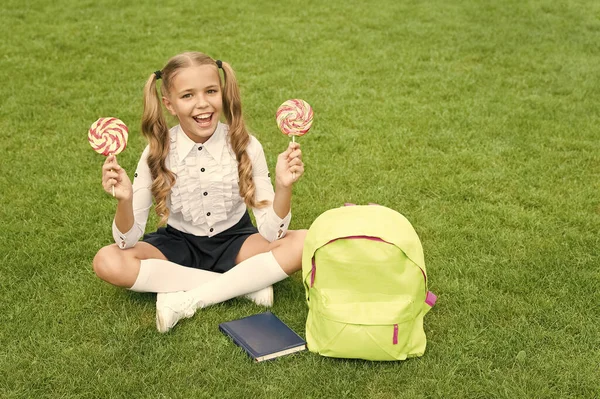 Okul üniformalı mutlu çocuk yeşil çimlerin üzerinde tatlı lolipoplar tutuyor, şekerler
