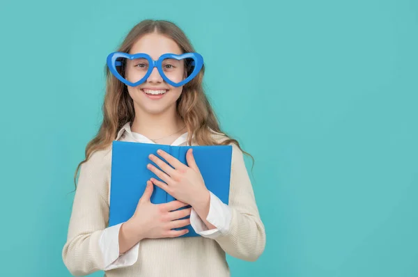Счастливый школьник держит школьную книгу в больших смешных очках в форме сердца синий фон — стоковое фото