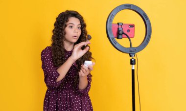 Şok olmuş kıvırcık saçlı çocuk blogcusu blog tutuş pudrası fırçası videosu yapıyor