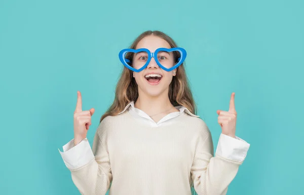 Glad flicka i stora roliga hjärtformade glasögon pekar fingrarna upp blå bakgrund, kopiera utrymme — Stockfoto