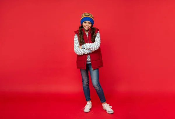 Pełna długość uśmiechniętego dziecka w dzianinie czapka zimowa i kamizelka puchowa na czerwonym tle, strój zimowy — Zdjęcie stockowe
