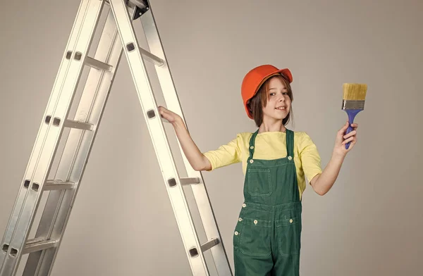 Девушка-работница в защитном шлеме и форме на лестнице с кистью, ремонт — стоковое фото