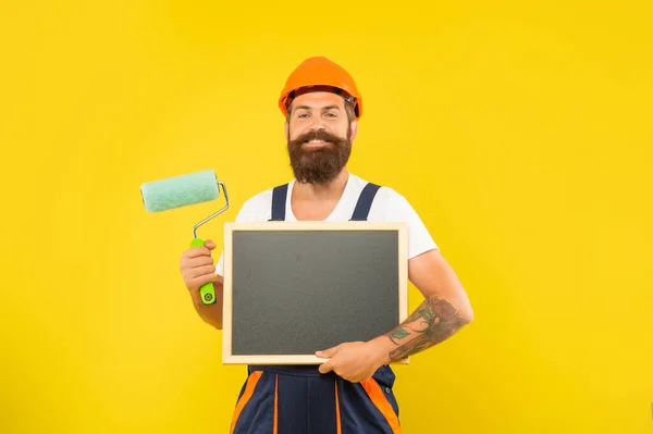 Lächeln Mann Dekorateur in Helm und Arbeitskleidung halten Farbroller und Tafel — Stockfoto