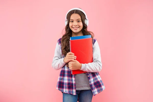 Happy kid listening music in earphones with workbook on pink background, education — ストック写真