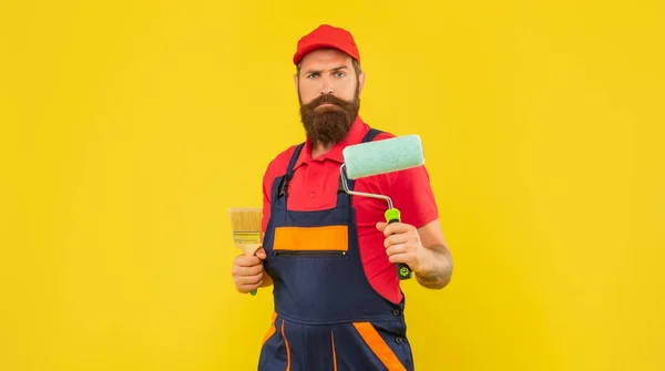 Бородатий чоловік художник в робочому одязі тримає фарбувальний валик і пензлик на жовтому тлі — стокове фото