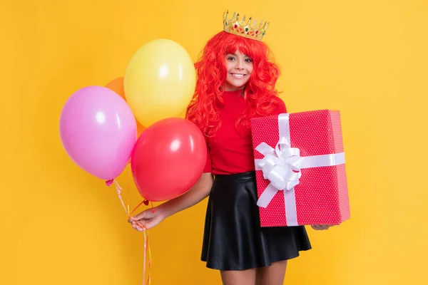 Happy girl in crown with present box and party balloon on yellow background — стоковое фото