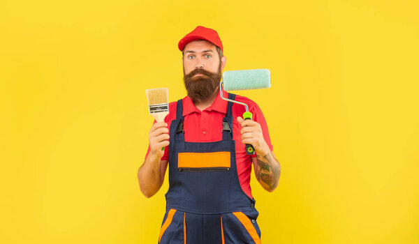 bearded puzzled man painter in work clothes hold paint roller and brush on yellow background
