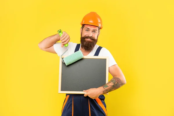 Positive man decorator in helmet and work clothes hold paint roller and blackboard with copy space — Stock Photo, Image