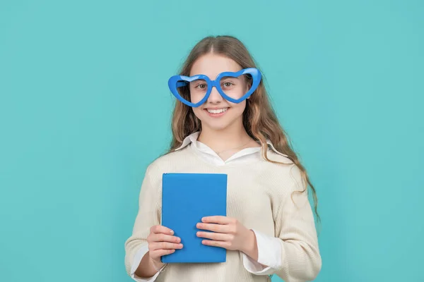Glückliches Kind zurück in die Schule mit Buch trägt große lustige herzförmige Brille blauen Hintergrund — Stockfoto