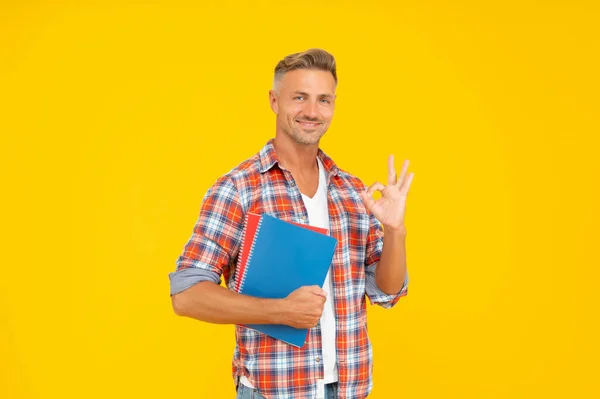 Heureux guy étudiant montrant OK anneau geste tenant livres jaune fond, université — Photo