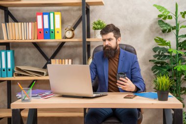 Kendine güvenen sakallı patron internette laptopta çalışıyor. Kahveyi ofiste tutuyor, e-postayı kontrol ediyor.