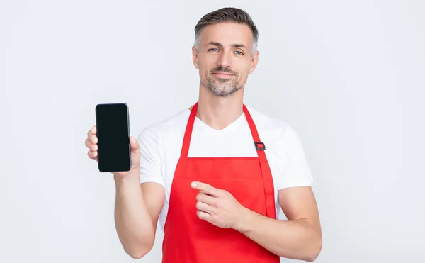 Cheerful mature man in apron presenting smartphone with copy space. point finger — Foto Stock