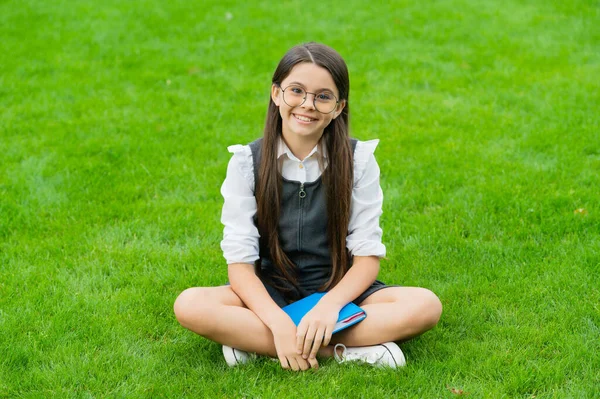 Smiling child in glasses sitting on green grass with book — 스톡 사진