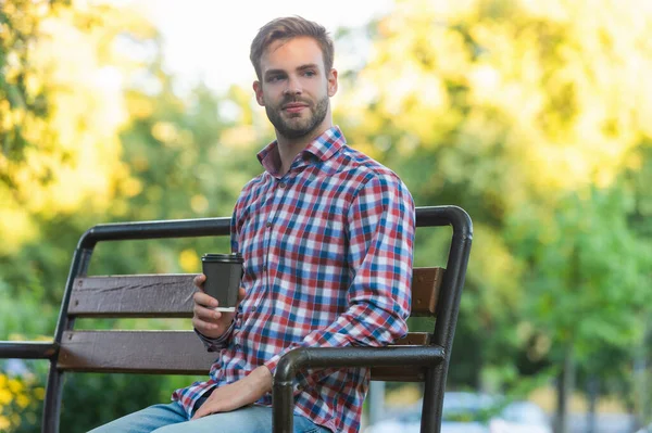 Smiling young man in checkered shirt with coffee cup on park bench — Foto de Stock