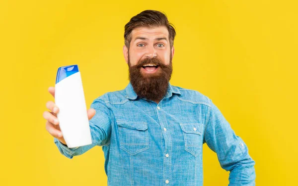 Изумленный жестокий бородатый мужчина держит бутылку шампуня на желтом фоне, уход за волосами — стоковое фото
