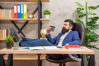 Tired man in suit relaxing with legs on desk during coffee break in office, relaxation