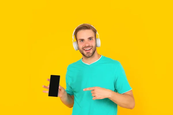Happy young man listening to music in headphones pointing finger at mobile phone, smartphone — 스톡 사진