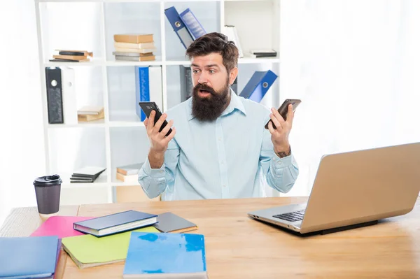 Shocked man with jaw dropped holding smartphones in each hand at office desk, mobile communication — Foto de Stock