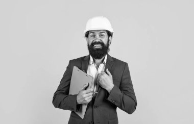 Takım elbiseli ve miğferli bir adam yan tarafa bakıyor ve sarı arka planda bir klasör tutuyor.