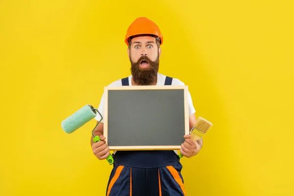 Shocked man renovator in helmet and work clothes hold paint roller brush and blackboard — Stockfoto