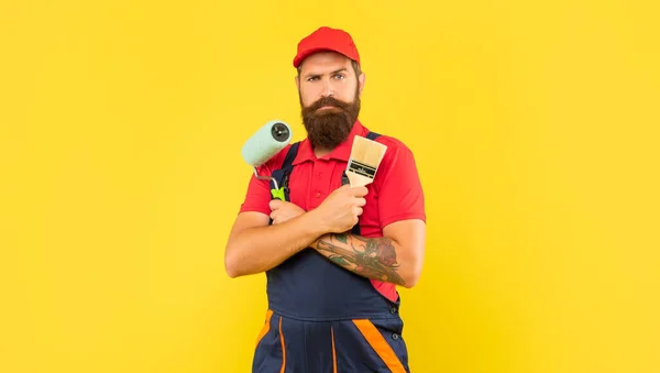 Pria berjanggut dalam pakaian kerja memegang rol cat dan kuas pada latar belakang kuning — Stok Foto