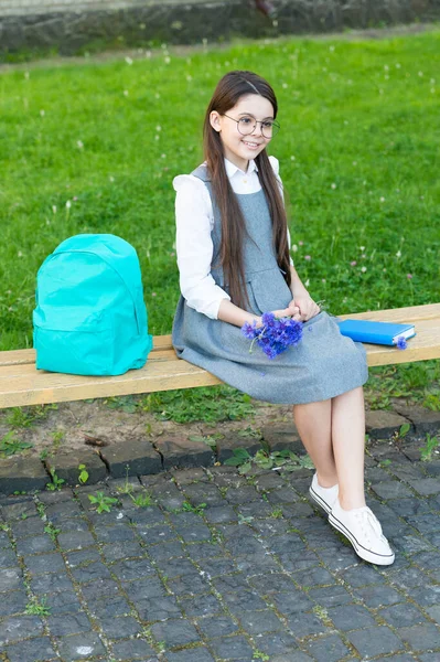 Niño feliz en gafas relajarse en el banco de la escuela con flores y mochila. día del conocimiento —  Fotos de Stock