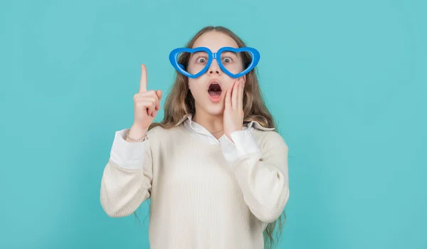 Překvapené dítě v legračních brýlích ve tvaru srdce, držící prst zvednutý na modrém pozadí, eureka — Stock fotografie