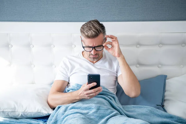 Mature man in bed reading phone wearing eyeglasses —  Fotos de Stock