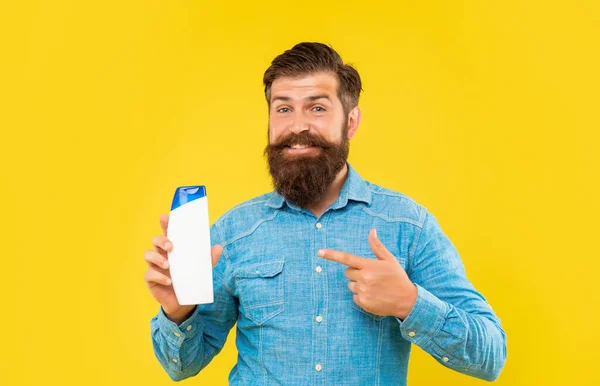 Счастливый жестокий бородатый мужчина указывать пальцем на бутылку кондиционера для волос на желтом фоне, уход за волосами — стоковое фото