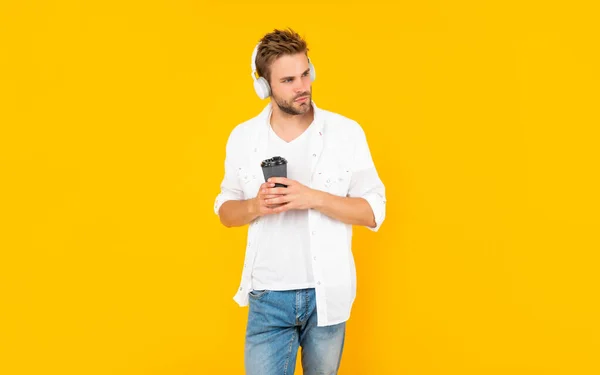 Hombre en camiseta blanca usar auriculares sostienen taza de café sobre fondo amarillo, dispositivo moderno — Foto de Stock