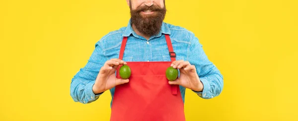 Happy man crop view in red apron holding fresh limes citrus fruits yellow background, greengrocers — Zdjęcie stockowe
