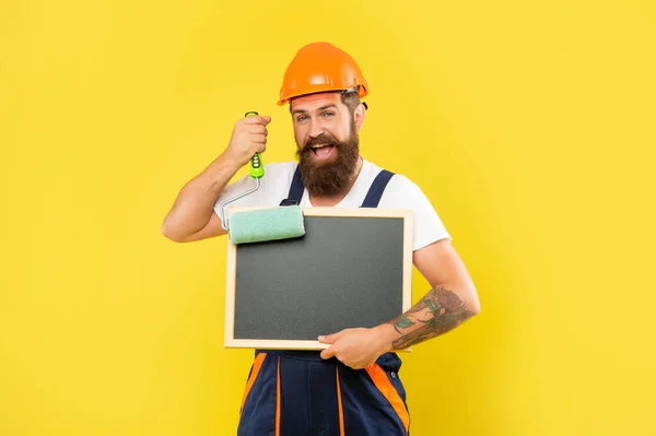 Cheerful bearded man in helmet and work clothes hold paint roller and blackboard — Stockfoto