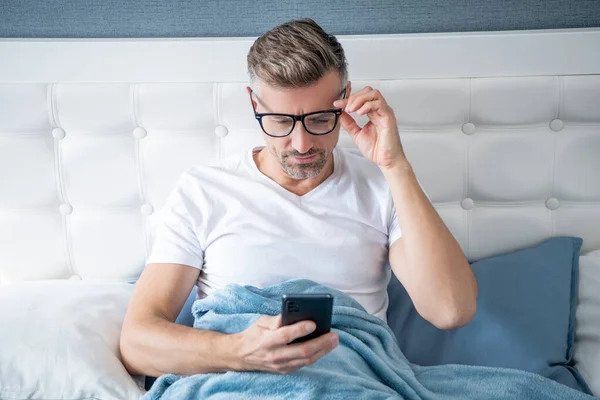 Mature man in bed chatting on phone wearing eyeglasses —  Fotos de Stock