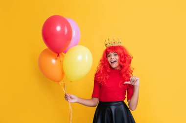 çocuk sarı arka planda helyum balonuyla taç içinde gülümsüyor