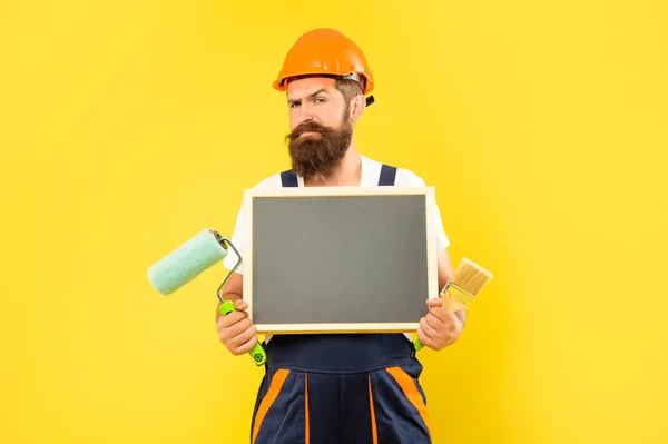 Man painter in helmet and work clothes hold paint roller brush and blackboard with copy space — Stockfoto