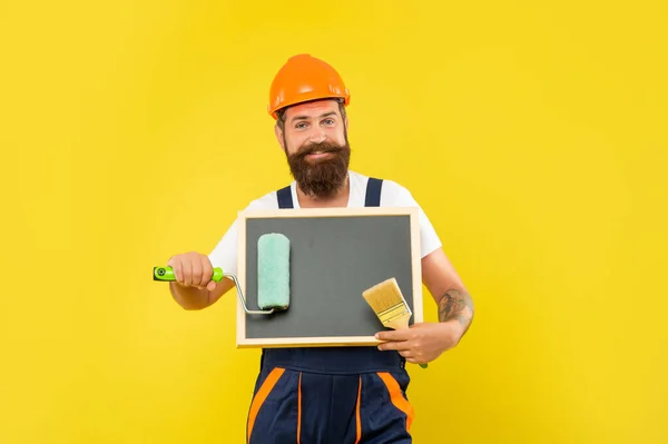 Fröhlicher Mann in Helm und Arbeitskleidung hält Pinsel und Tafel in der Hand — Stockfoto