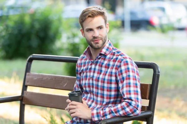 Cheerful young man in checkered shirt with coffee cup on park bench — Stok fotoğraf