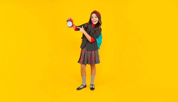 Amazed teen girl with school bag pointing finger on alarm clock on yellow background, time. — Foto de Stock