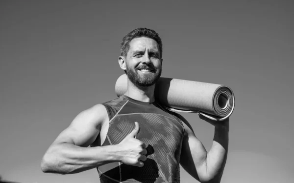 Pilates trener człowiek z maty do jogi pokazując kciuk w górę na tle nieba, sukces sportowy — Zdjęcie stockowe