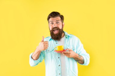 Kahve fincanı tutan sakallı hippi şaşırdı sarı arka planda baş parmağını gösteriyor, kahve...