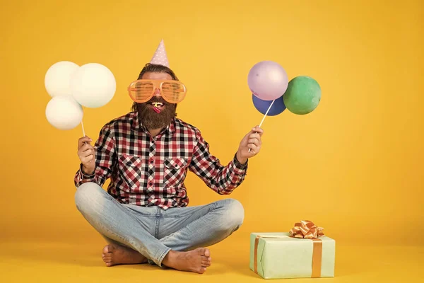 Glücklich schöner bärtiger Kerl mit unrasiertem Gesicht und stylischer Frisur trägt lässige Kleidung mit Geschenk und Party-Luftballons, Geburtstagsurlaub — Stockfoto