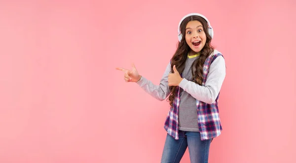 Έκπληκτος παιδί ακούγοντας μουσική σε ακουστικά δείχνουν δάχτυλο σε ροζ φόντο με αντίγραφο χώρο, τεχνολογία — Φωτογραφία Αρχείου