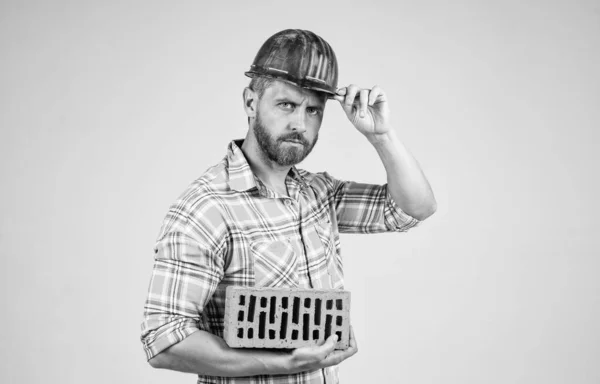 Ο άνθρωπος χτίζει σπίτι. ώριμος οικοδόμος με πουκάμισο. αξύριστος άνθρωπος στο εργοτάξιο. Όμορφος οικοδόμος με σκληρό καπέλο. Πρωτομαγιά. Ο τύπος φοράει κράνος ασφαλείας και κρατάει τούβλο. μηχανικός στη δουλειά. ανδρικός τρόπος ζωής — Φωτογραφία Αρχείου