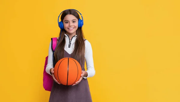 Vrolijk kind in hoofdtelefoon met school rugzak en basketbal op gele achtergrond — Stockfoto